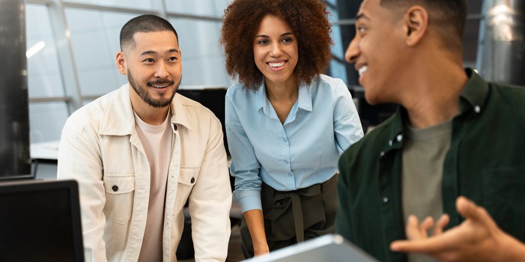 Três jovens em frente ao computador conversando entre si e sorrindo.